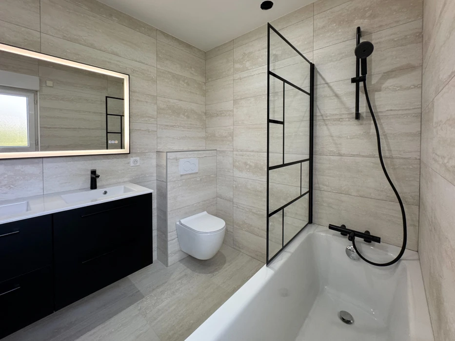 Salle de bain moderne avec baignoire d'angle, douche à l'italienne séparée par cloison vitrée noire, meuble vasque suspendu noir, toilettes suspendues et miroir éclairé